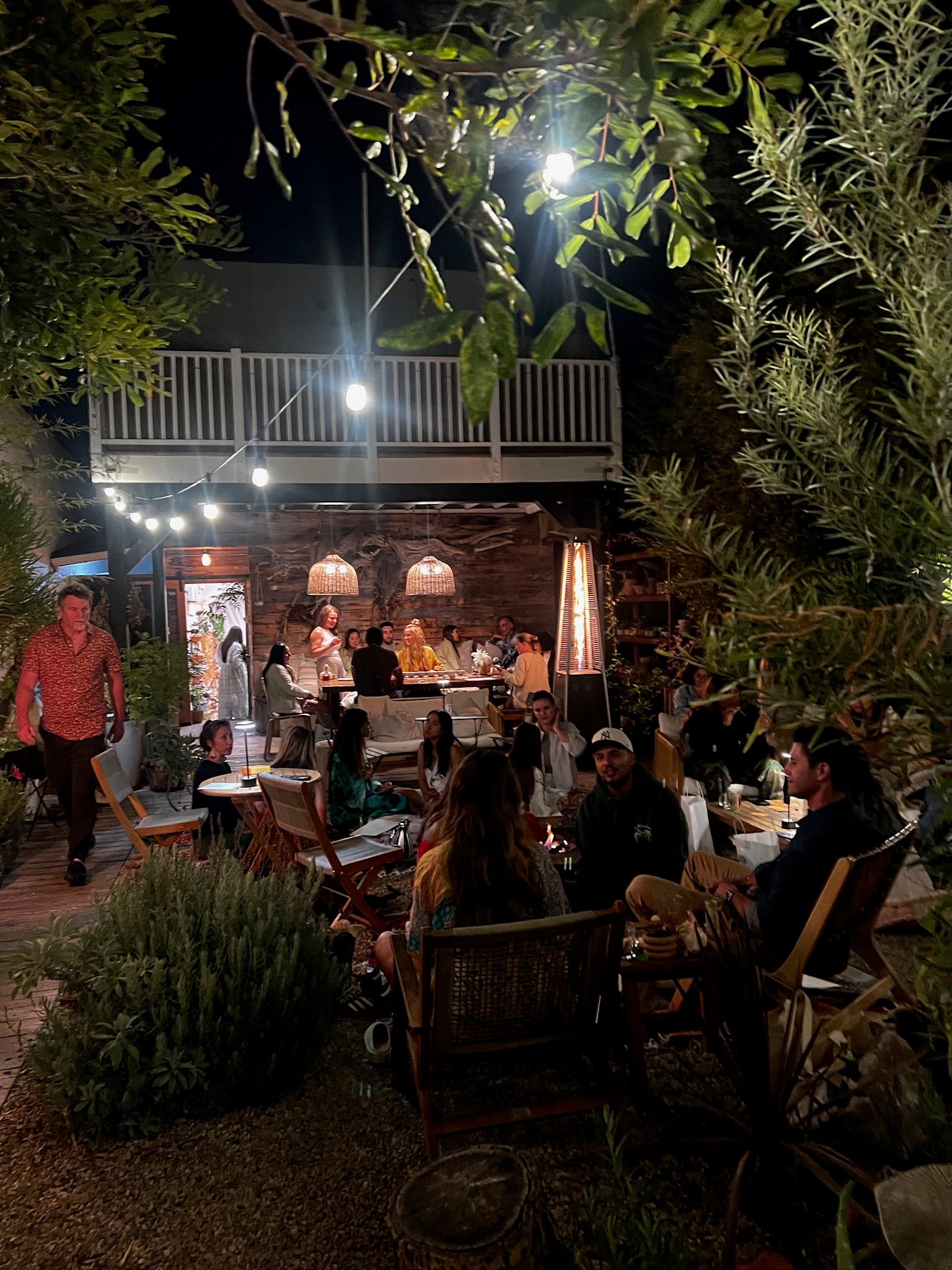 Outdoor evening gathering with people seated at tables under string lights and a building in the background.