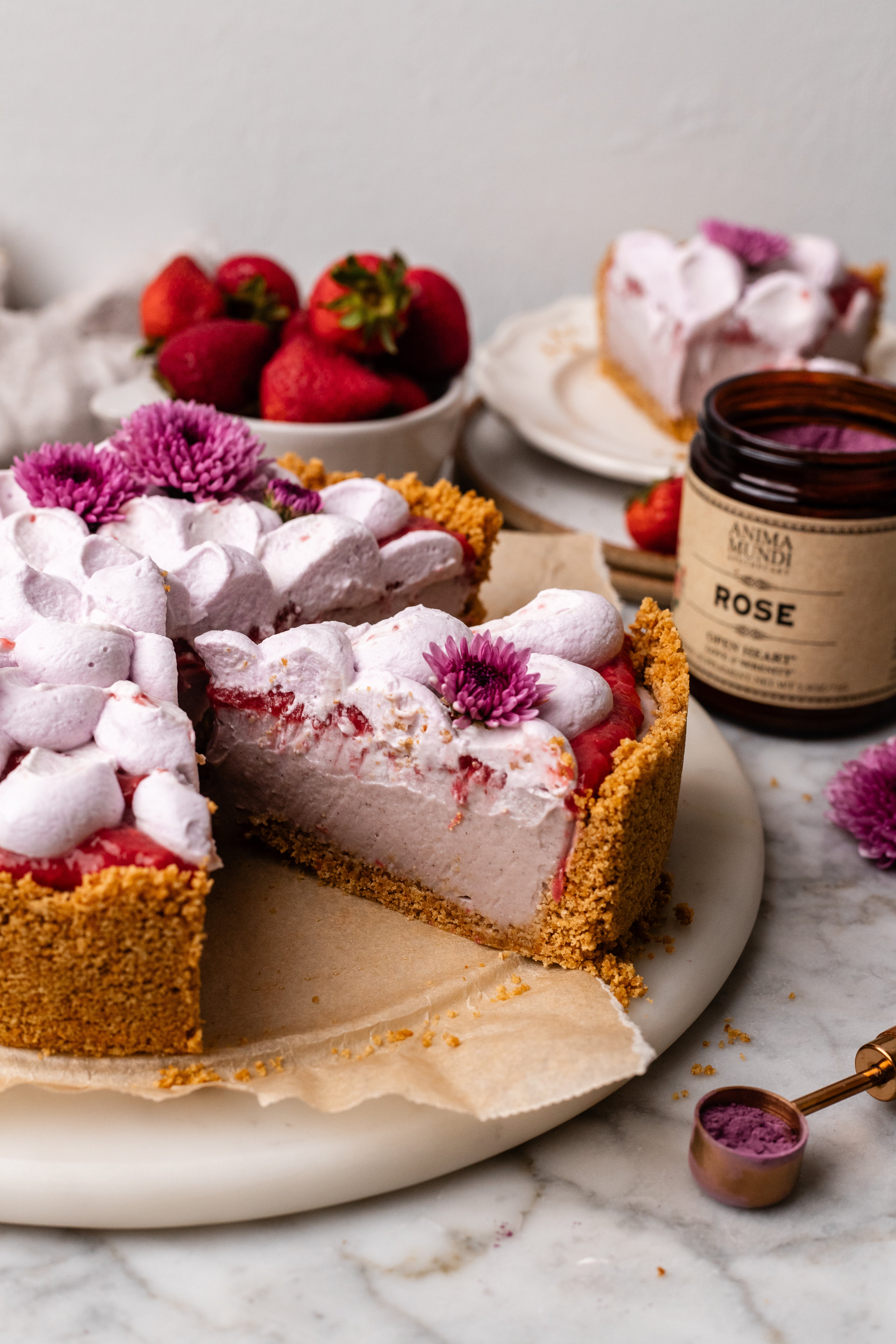 Cheesecake with pink whipped cream and strawberries on a marble surface with a jar of rose extract.