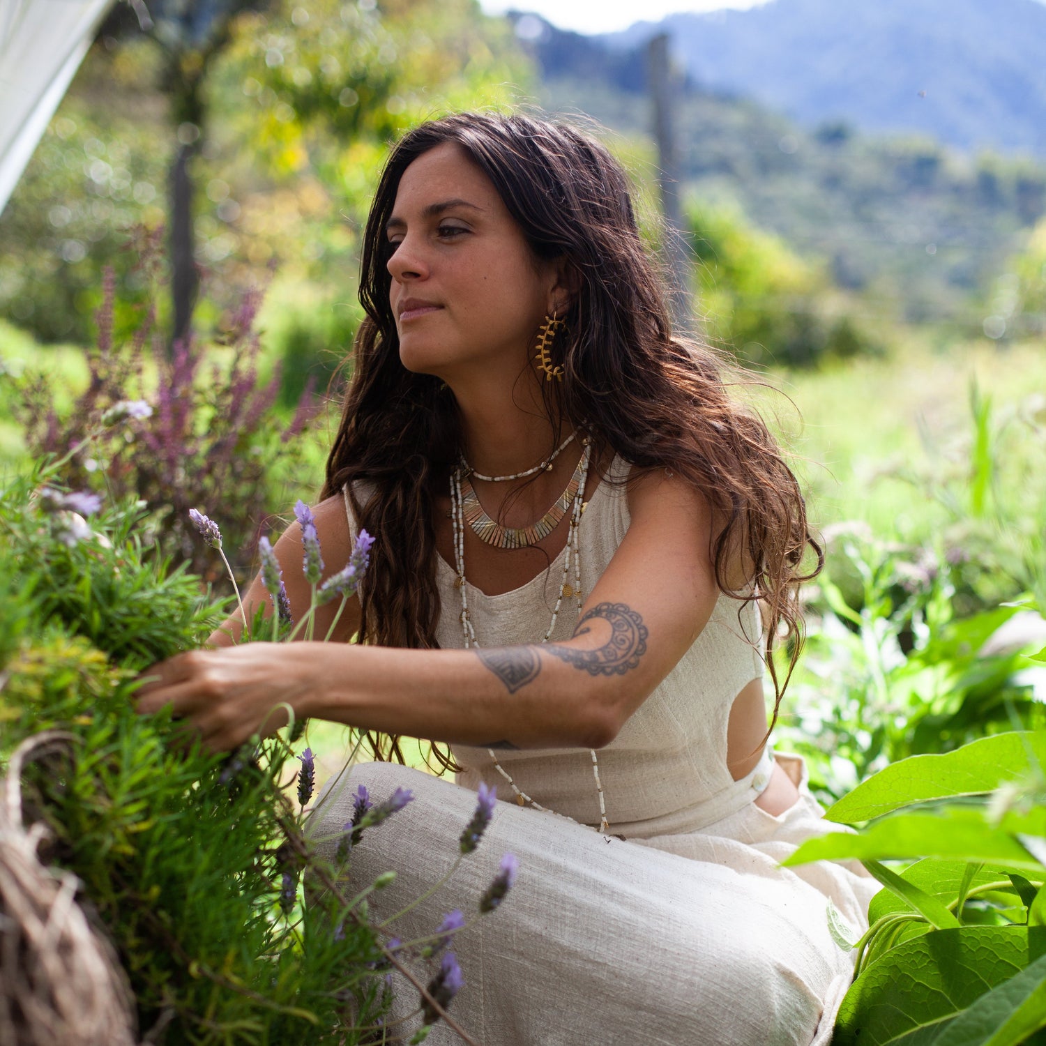 A woman with long dark hair crouches in a lush garden, wearing a sleeveless cream linen dress and layered gold necklaces. She is surrounded by green plants and purple flowers, with mountains in the background.