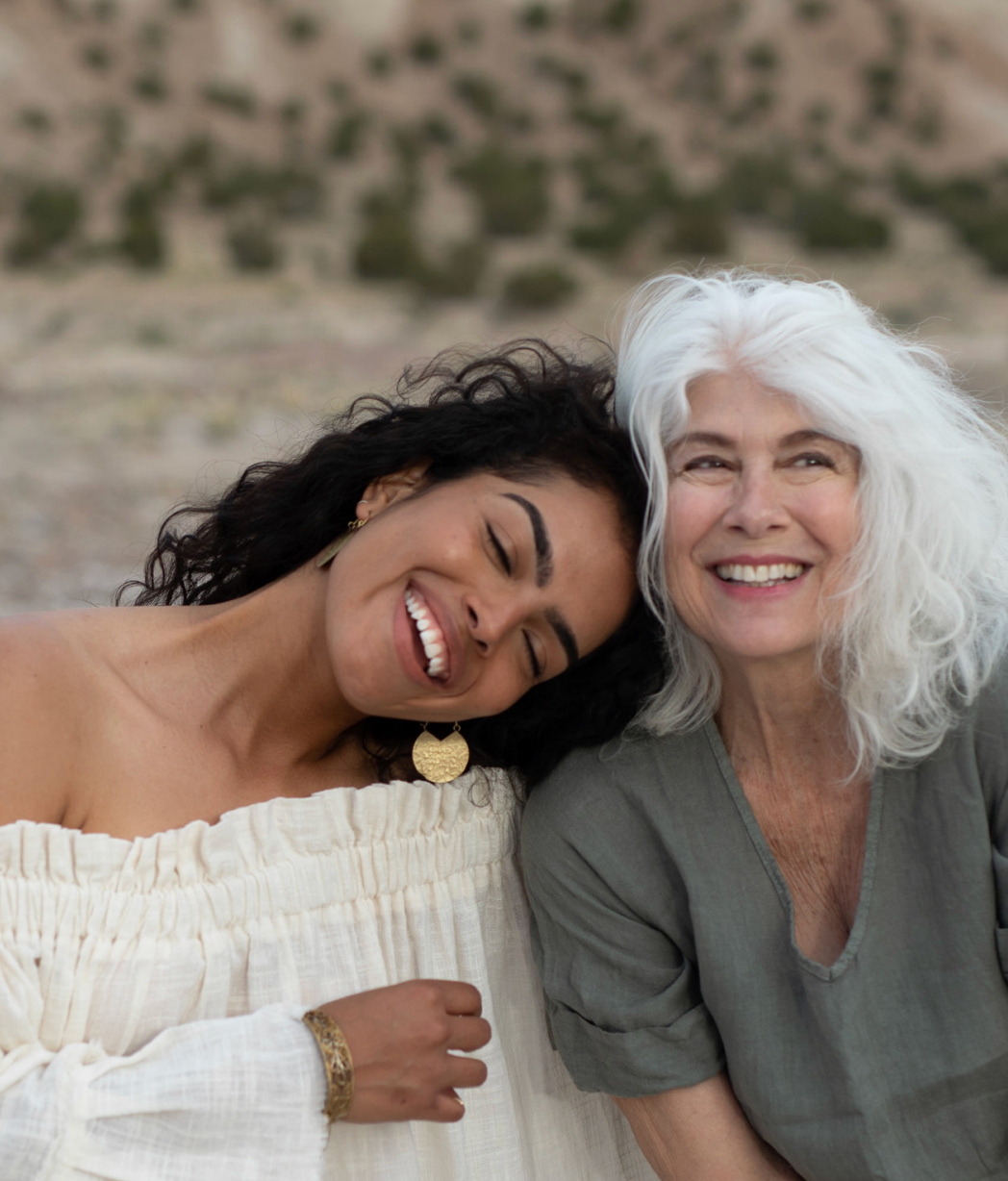 Two women smiling and embracing outdoors with a natural background