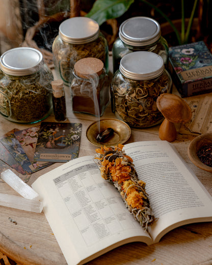 A styled tabletop arrangement featuring an open book with printed text, a sage smudge bundle, several glass jars containing dried herbs or spices, a small smoky incense holder, tarot cards, a wooden mushroom-shaped object, and a crystal cluster on a wooden surface.