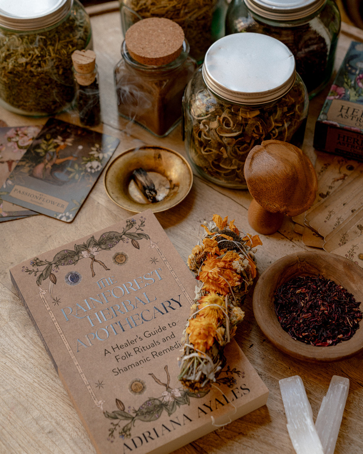 The Painforest Herbal Apothecary book by Adriana Jales lies on a wooden table surrounded by herbalist props including glass jars of dried herbs, a wooden mushroom-shaped tool, a sage bundle, a bowl of red botanicals, tarot cards, and crystal points.