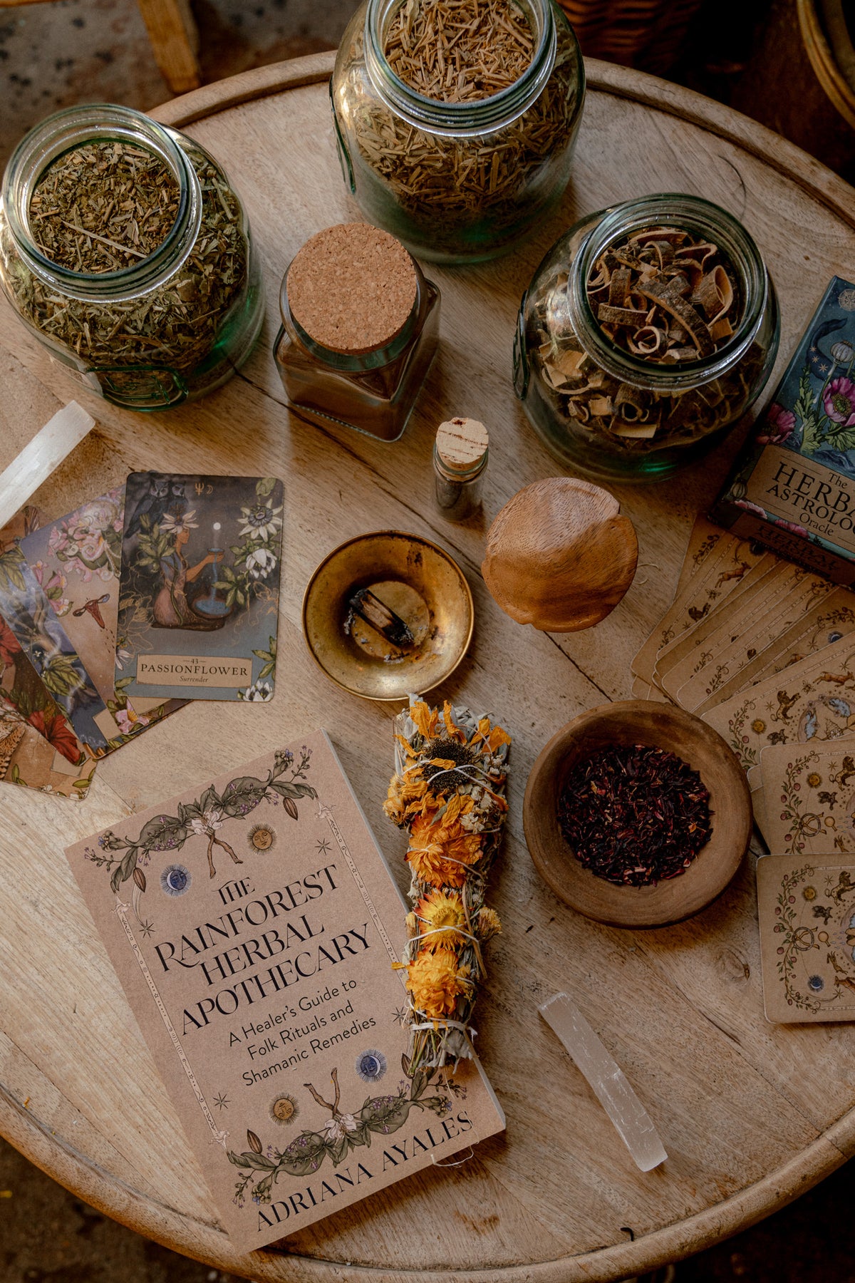 A styled flat-lay of a book titled 'The Panforest Herbal Apothecary' by Adriana Byles, surrounded by glass jars of dried herbs, a sage bundle, crystals, cards, and small wooden bowls on a round wooden table.
