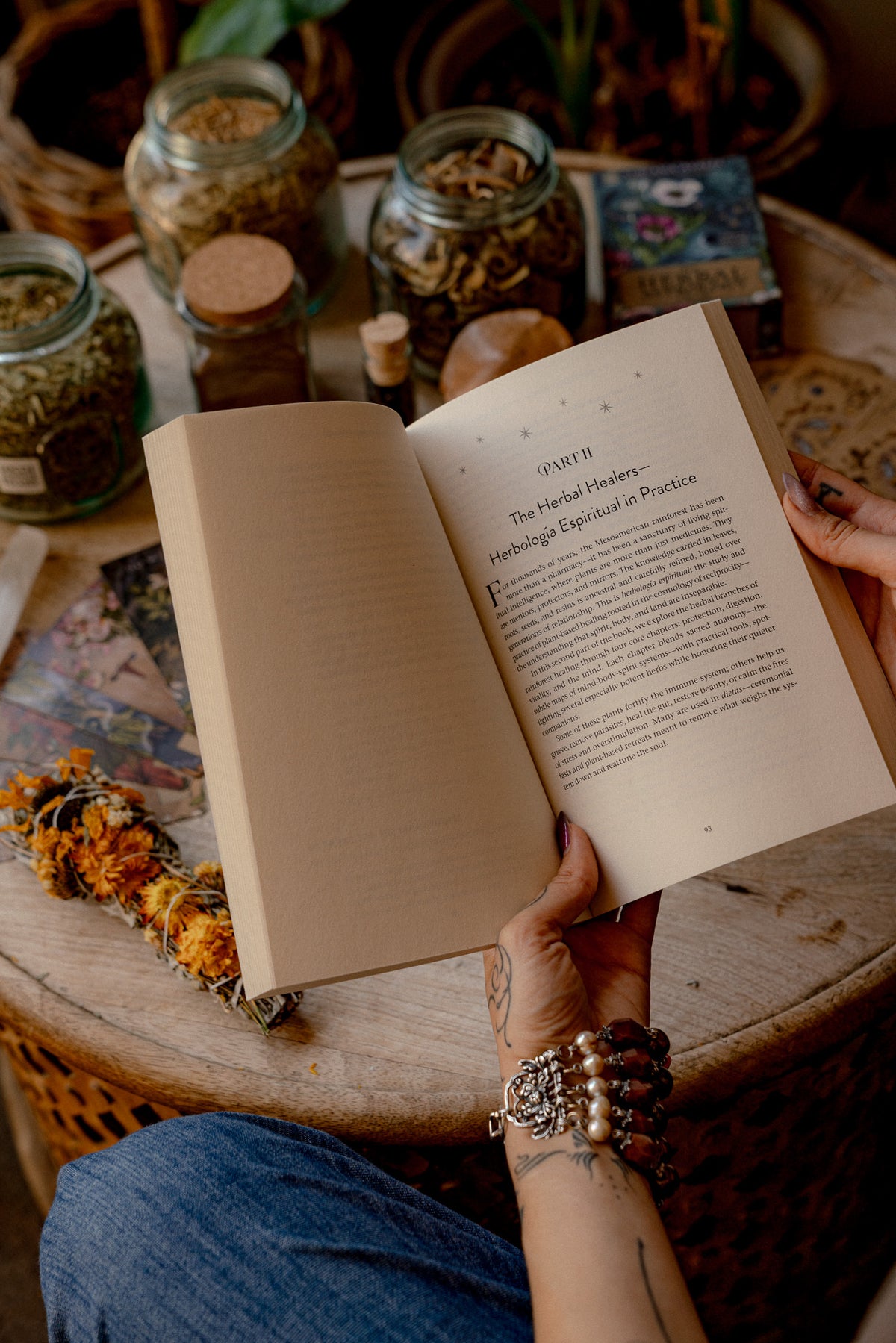 A person holding an open book with printed text, photographed from above on a rustic wooden table surrounded by glass jars of dried herbs and botanical items.