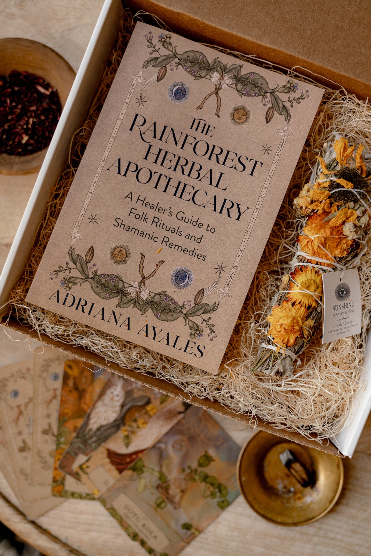 An open gift box containing a book titled "The Painforest Herbal Apothecary" by Adriana Ayales, surrounded by herbal remedies including dried flowers, cards, and a brass dish on a rustic wooden surface.