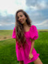 A woman with long wavy brown hair wearing a bright pink puff-sleeve mini dress, standing in a grassy field under a cloudy blue sky.