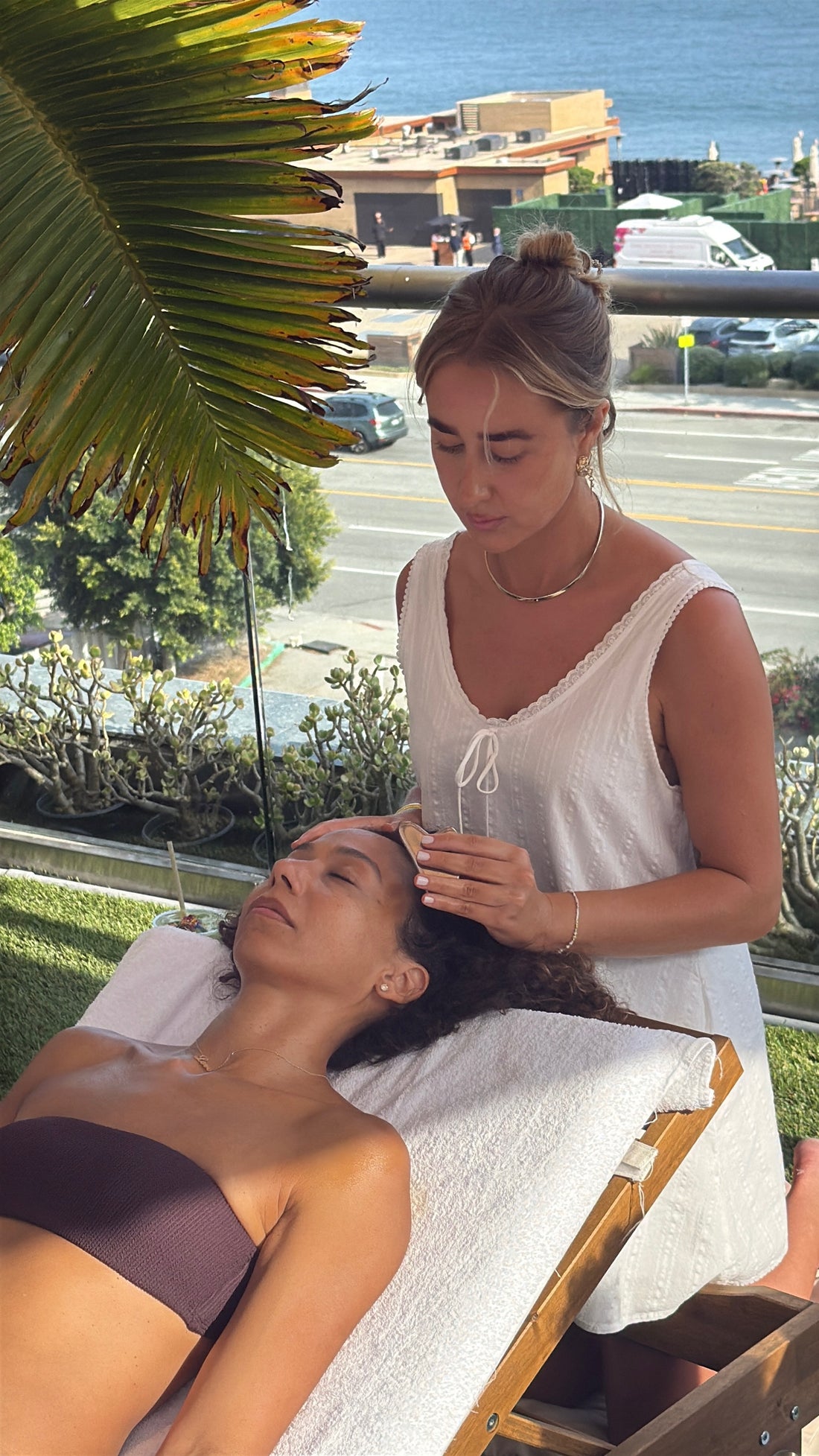 A woman in a white sleeveless top massages the head of another woman lying on a white lounge chair outdoors on a balcony with a view of the ocean and palm leaves.