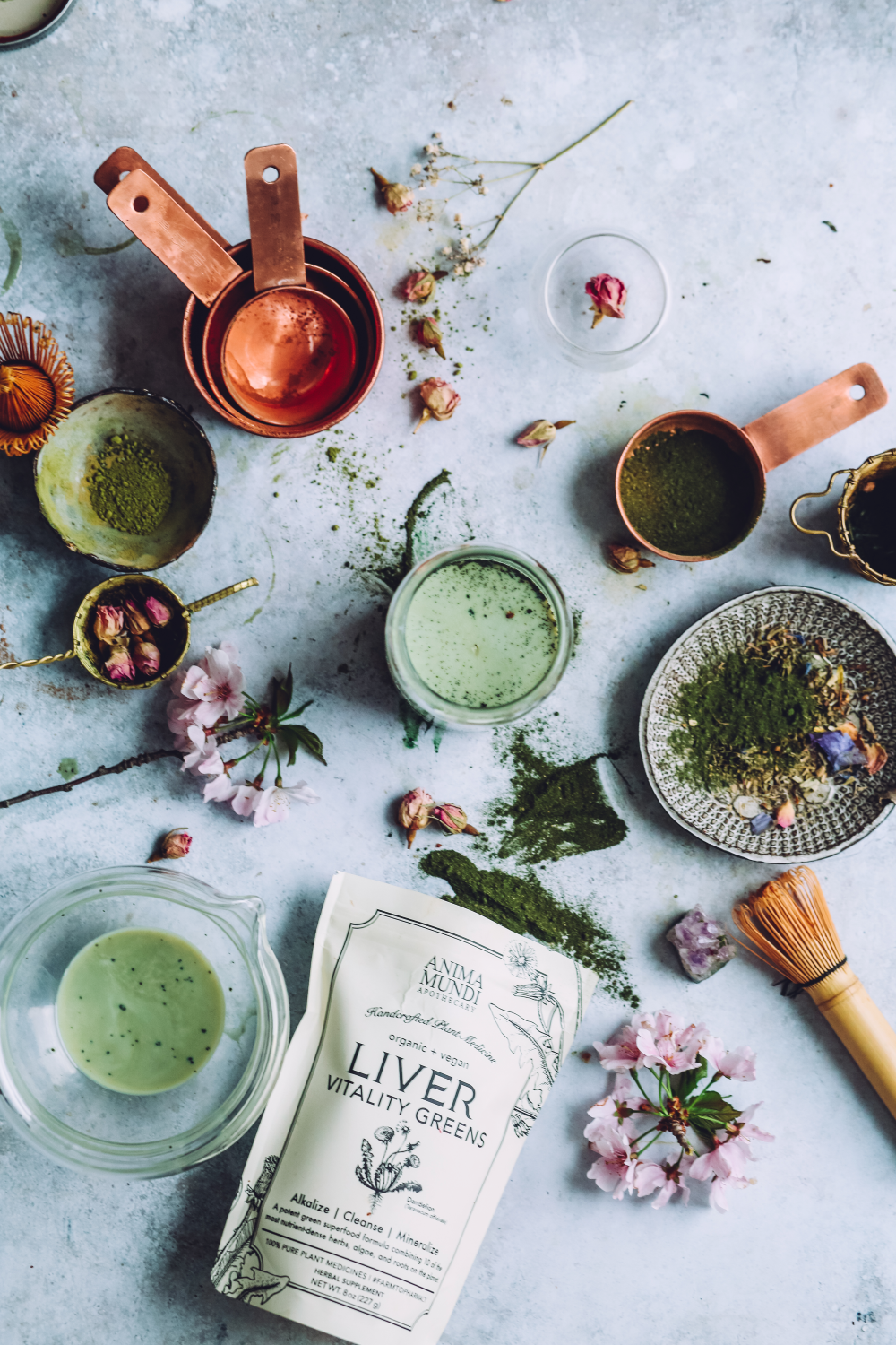 Green powder in measuring cups, glasses, and a package labeled 'Liver Vitality Greens' on a light blue surface with flowers and herbs.