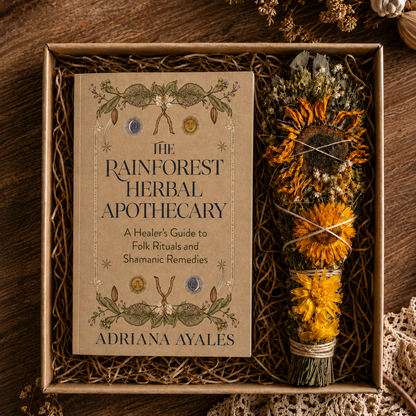 A boxed book titled "The Rainforest Herbal Apothecary" by Adriana Ayales, placed on a rustic wooden surface with a dried flower bundle beside it. The scene is styled with natural props and a warm earthy background.
