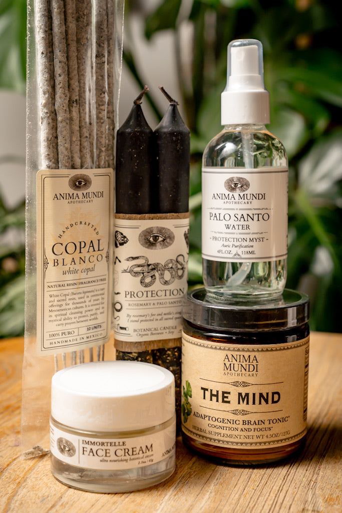Collection of Anima Mundi products including candles, incense, and face cream on a wooden surface with greenery in the background.